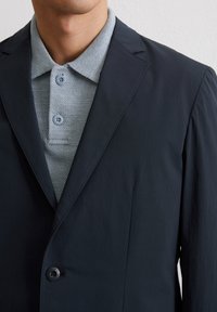 Granatowa marynarka o gładkiej fakturze, z pojedynczym zapięciem na guzik i spiczastymi klapami. Pod spodem jasnoszara, teksturowana koszulka polo.