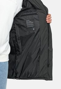 Veste en duvet gris foncé, extérieure en nylon, poche intérieure avec zip, texture douce, étiquette avec instructions d'entretien visible.