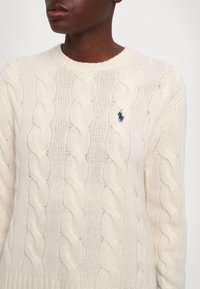 Personne portant un pull en maille torsadée crème avec un logo de cheval brodé bleu marine sur le côté gauche de la poitrine, photographiée du cou jusqu'au milieu du torse.