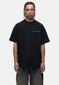 Black cotton oversized t-shirt with short sleeves, round neckline, and white text logo on the left chest area; smooth texture.