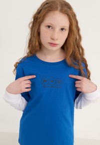 Blue t-shirt featuring a round logo with "NYC" and "NewYouthCulture" in black. Long white sleeves visible at the cuffs. Smooth fabric texture.