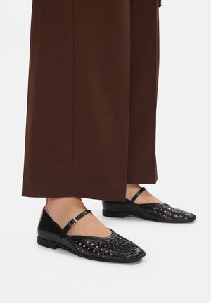 Czarne płaskie buty z plecionej tkaniny z noskiem w szpic i paskiem wokół kostki, o teksturowanej powierzchni. Stylizowane z szerokimi brązowymi spodniami.