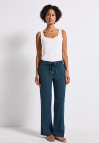 Pantaloni larghi color teal scuro con una vita elasticizzata e cordino regolabile, abbinati a una maglietta senza maniche bianca. Tessuto di texture liscia.