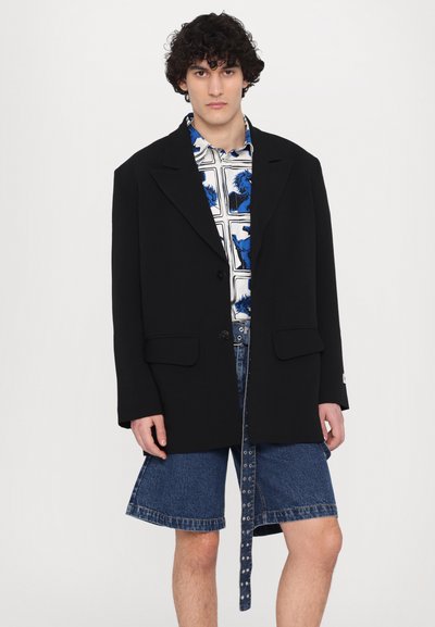 Black oversized blazer with notched lapels, paired with a blue graphic shirt and denim shorts featuring a decorative belt and snap closures.