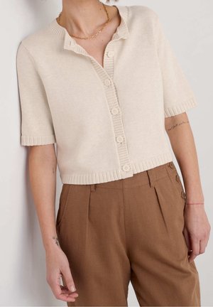Femme portant un cardigan crème à manches courtes boutonné et un pantalon marron taille haute avec des boutons sur les côtés, sur un fond uni.