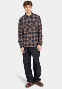 Geruite flanellen blouse in marineblauw en oranje tinten, met een knoopsluiting, twee borstzakken en een ontspannen pasvorm. Gecombineerd met donkere denim broek.