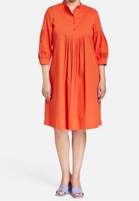 Vestito di cotone arancione con colletto a punta, maniche a tre quarti a sbuffo, pieghe centrali e dettagli con bottoni. Abbinato a sandali a scivolo viola.
