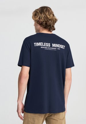 T-shirt in cotone blu navy con la scritta "TIMELESS MINDSET" sul retro in bianco. Collo giro standard e maniche corte.