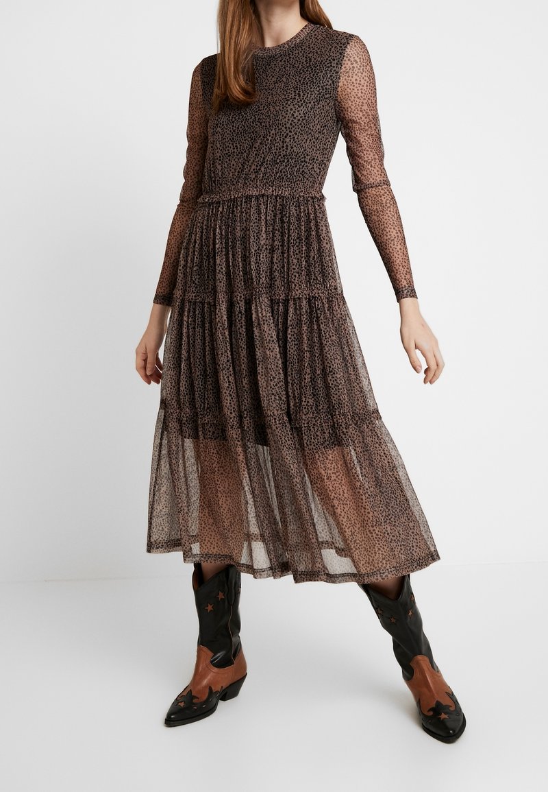 Femme portant une longue robe marron transparente à pois avec une jupe à volants et des manches longues, associée à des bottes de cowboy noires et marron avec des découpes en forme d'étoile.