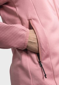 Giacca rosa testurizzata con tasca con zip. Presenta dettagli a coste e accenti in corda nera. Primo piano della mano che si inserisce nella tasca.