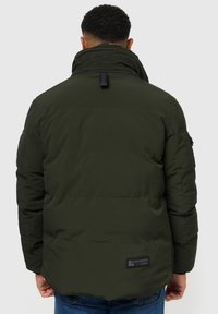 Giubbotto imbottito verde oliva con colletto alto, cuciture visibili e una patch con logo sul retro. Presenta una chiusura con zip e tasche laterali.