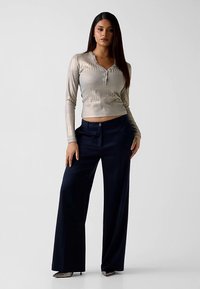 Haut à manches longues gris clair brillant avec texture côtelée et détails de boutons, associé à un pantalon bleu marine taille haute à jambes larges.