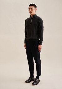 Maglione grigio scuro con zip e collo alto, abbinato a pantaloni blu navy e scarpe nere con lacci, indossato da una modella su sfondo neutro.