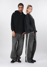 Δύο άτομα στέκονται δίπλα-δίπλα, φορώντας φούτερ με κουκούλα και φαρδιά γκρι τζιν. Τα τζιν έχουν λευκές πλάγιες ρίγες· τα υφάσματα φαίνονται μαλακά και με υφή.