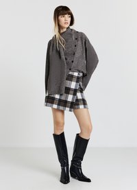 Giacca grigia con texture e bottoni, gonna a quadri in nero, bianco e beige, abbinata a stivali neri in pelle fino al ginocchio.