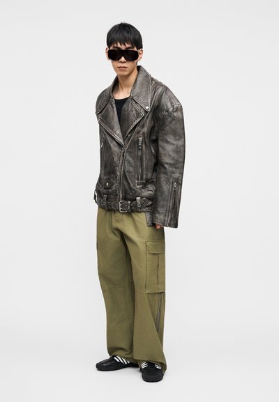 Μαύρο δερμάτινο μπουφάν μοτοσικλετιστή με υφή, ασημί εξαρτήματα, υπερμεγέθη παντελόνια cargo σε ελιά πράσινο και μαύρα γυαλιά ηλίου.