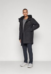 Svart vattentät parkas med huva, flera fickor och dragkedja framtill. Bärs över en grå t-shirt och mörka byxor. Texturerat tyg.