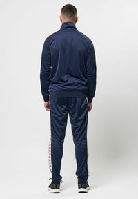 Trening navy, realizat dintr-un material neted, având un guler înalt, dungi laterale cu accente de logo și manșete elastice atât la jachetă, cât și la pantaloni.