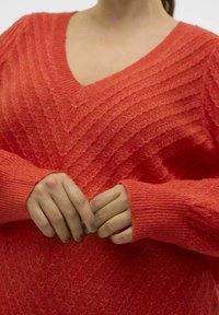 Červený pletený svetr s véčkovým výstřihem, texturovaným zigzag vzorem a žebrovanými manžetami. Materiál vypadá měkce a mírně melírovaně.