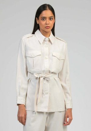 Femme portant une veste-chemise beige boutonnée avec des poches poitrine et une ceinture nouée à la taille, debout devant un fond gris uni.