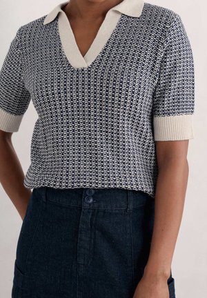 Person wearing a navy and cream textured knit short-sleeve polo shirt tucked into high-waisted dark denim pants with buttons.