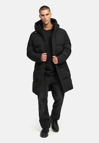 Zwarte pufferjas met een capuchon, rits aan de voorkant en een gequiltde textuur. Draagt over een zwarte longsleeve en zwarte broeken, aangevuld met laarzen.