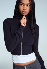 Maglione blu navy con zip, colletto alto, materiale testurizzato e maniche lunghe. Indossato sopra una canotta bianca, abbinato a pantaloni grigi.