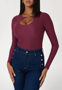 Ilga rankovė, bordo spalvos kūno kostiumas su plyšiu kaklo srityje, derinamas su aukštu liemeniu džinsais, kuriuose yra auksinių mygtukų akcentų.
