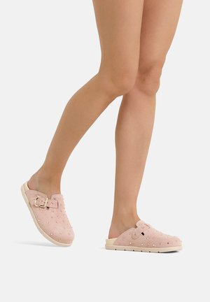 Persona che cammina a piedi nudi indossando zoccoli rosa pallido slip-on con borchie decorative e fibbia su suola bianca su sfondo bianco.