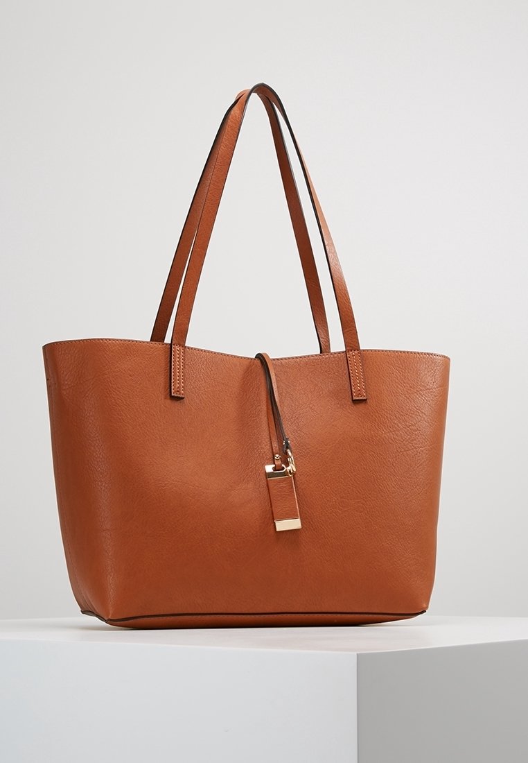 Sac tote en cuir marron avec doubles bretelles et une petite étiquette suspendue à accents dorés, posé sur un socle blanc sur un fond uni.
