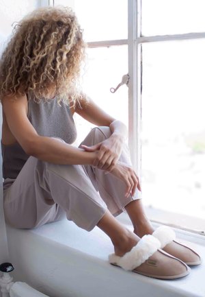 Zapatillas desenfocadas de un suave color beige claro con un borde de piel sintética blanca, que presentan un material suave y suela plana, colocadas sobre un borde blanco cerca de una ventana.