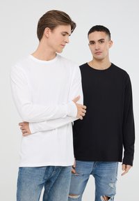 Due modelle indossano camicie a maniche lunghe: una bianca e una nera, entrambe con scollatura tonda. Le modelle indossano jeans azzurri strappati.