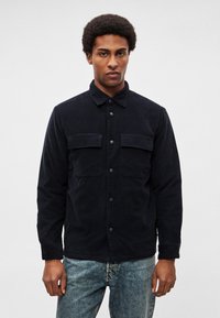 Jakna tamnoplave boje s gumbima, izrađena od mekanog tkanja. Ima dva džepa na prsima i šiljati ovratnik. Nošena je uz svijetloplave traperice.