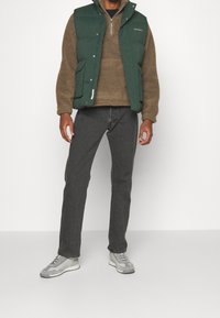 Grüne wattierte Weste mit Druckknöpfen, brauner Fleece-Pullover darunter, dunkelgraue Jeans und graue Sneakers mit Muster. Ein Mann steht vor einem weißen Hintergrund.