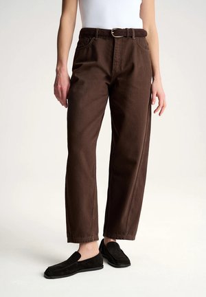 Osoba w brązowych spodniach z wysokim stanem i paskiem, białym bezrękawnym topie oraz czarnych wsuwanych butach, stojąca na jednolitym jasnym tle.