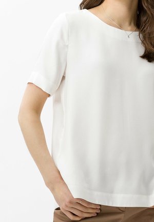 Woman wearing a plain white short-sleeve blouse, with a silver necklace and light brown pants, standing with one hand on her hip.