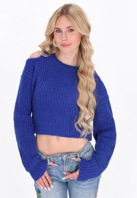 Maglione cropped in un blu vivace, caratterizzato da un motivo testurizzato, maniche larghe e design con spalle scoperte. Abbinato a jeans azzurri.