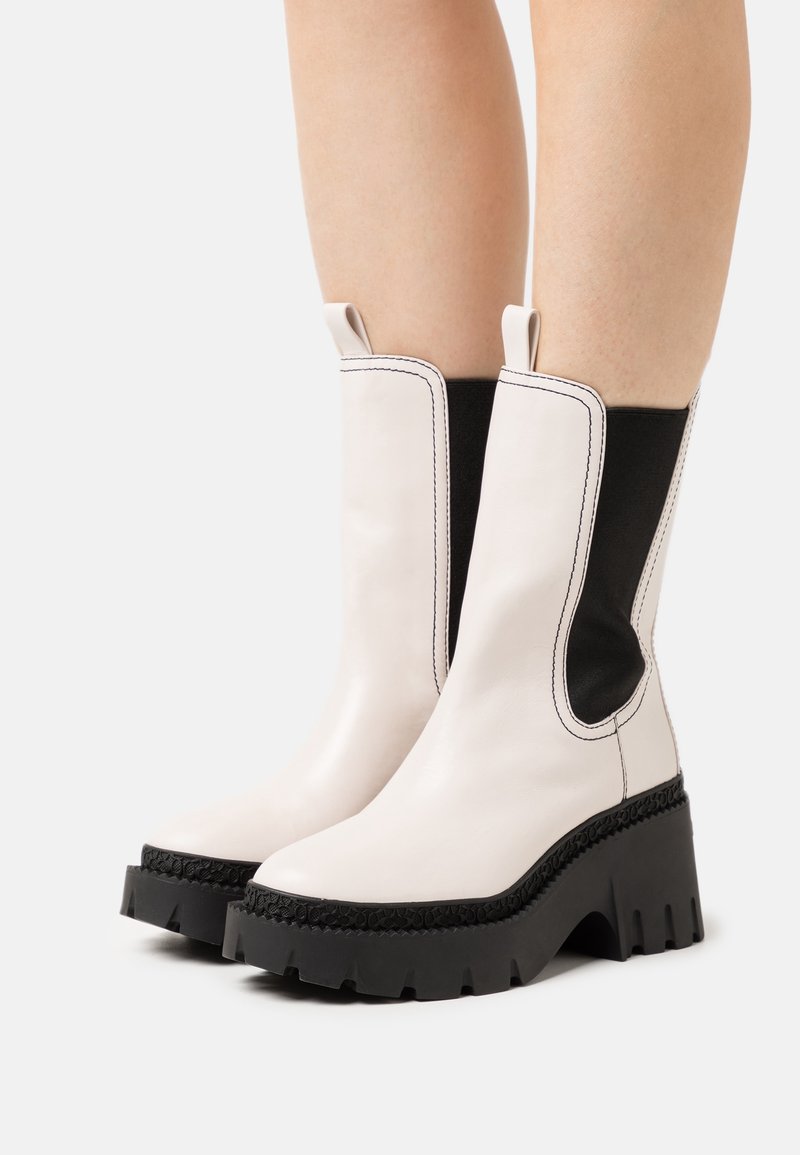 Bottes blanches mi-mollet avec semelles épaisses noires, panneaux latéraux élastiques noirs et languettes de tirage, portées sur des jambes nues sur un fond uni.