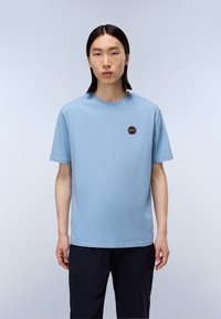 Světle modré bavlněné tričko s kulatým výstřihem a krátkými rukávy, s malou kulatou nášivkou na levé části hrudi.
