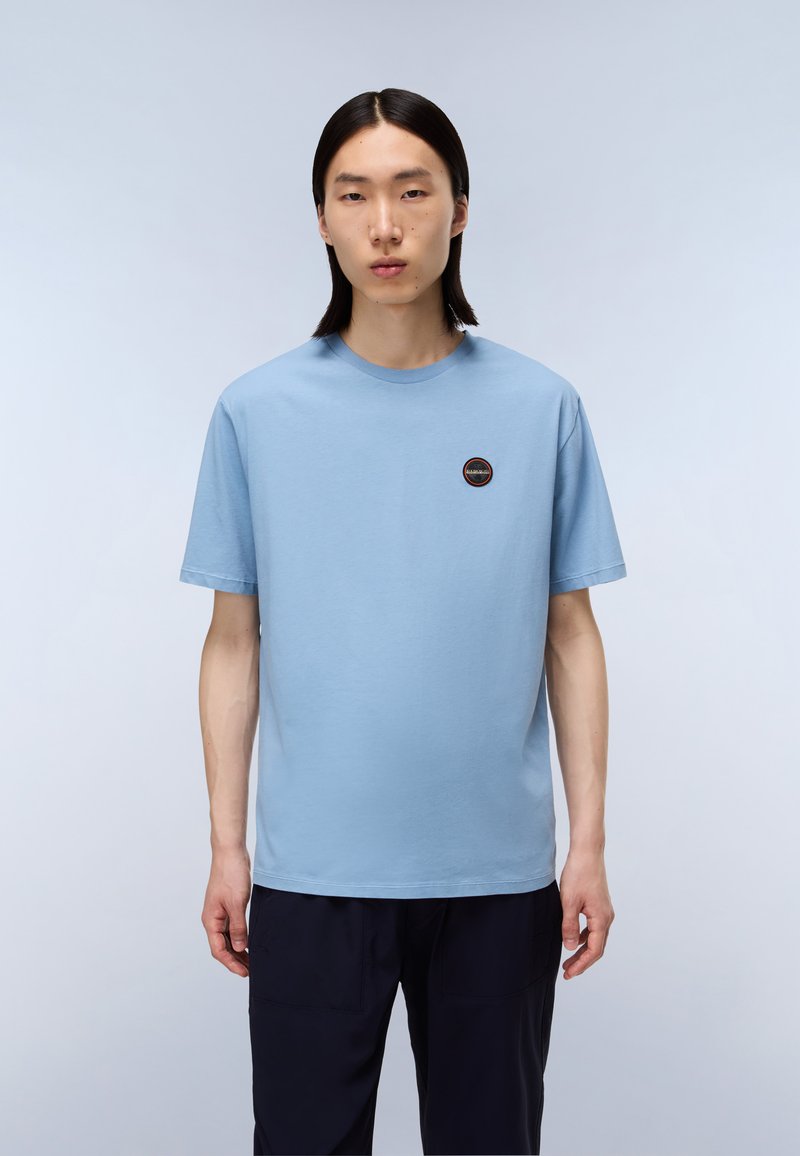 Světle modré bavlněné tričko s kulatým výstřihem a krátkými rukávy, s malou kulatou nášivkou na levé části hrudi.