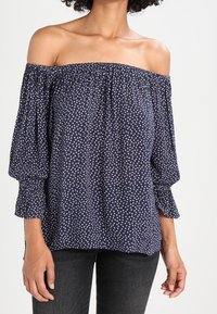 Kvinna som bär en marinblå off-shoulder-blus med vita prickar och svarta byxor, stående mot en ljus bakgrund.