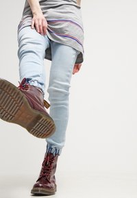 Jeans skinny azul claro con puños elásticos, combinados con botas de cuero burdeos con cordones. Camisa gris con rayas horizontales moradas y rojas visibles.
