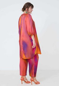 Zijden outfit met een lange, losse tuniek met levendige oranje, roze en paarse ombre-patronen, gecombineerd met bijpassende broeken en oranje hakken.