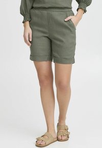 Olive green shorts made of lightweight fabric with rolled cuffs and side pockets, paired with beige sandals featuring large buckles.