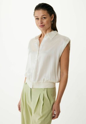 Light silk blouse with short, rolled sleeves, button front, and elasticized hem. Worn with light green pleated trousers. Neutral background.