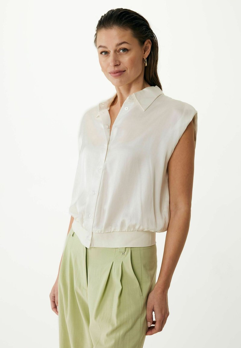 Blusa di seta leggera con maniche corte arrotolate, chiusura a bottoni e orlo elastico. Indossata con pantaloni plissettati di colore verde chiaro. Sfondo neutro.