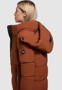 Chaqueta de plumas marrón con capucha, con un diseño acolchado, detalles en metal negro, bolsillos laterales con cremallera y logotipo en la manga.