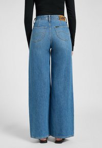 Bredde ben blå jeans laget av denim, med høy midje, baklommer og en brun lærlogo-patch på midjebåndet.