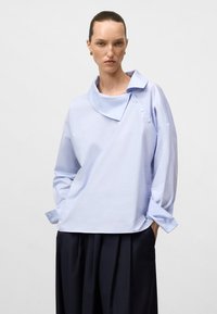 Jasnoniebieska, oversize bluzka z załamanym kołnierzykiem i trzema guzikami z boku, wykonana z gładkiego materiału, w komplecie z ciemnymi, plisowanymi spodniami.