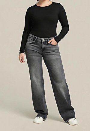 Femme portant un haut noir à manches longues, jean gris délavé à taille haute et jambes larges, baskets blanches, debout avec une main dans la poche.